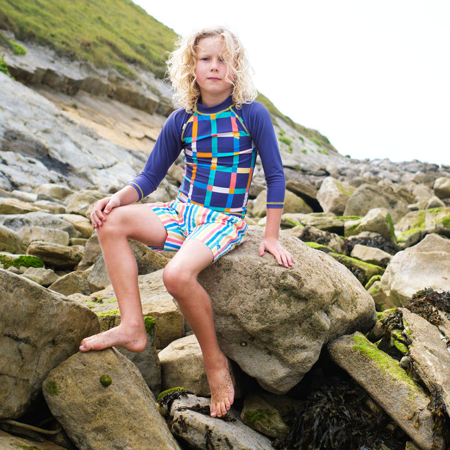 A boy sitting on rocks wearing UV Protective Swim Shorts Multi Stripe, rocks and hillside in background.